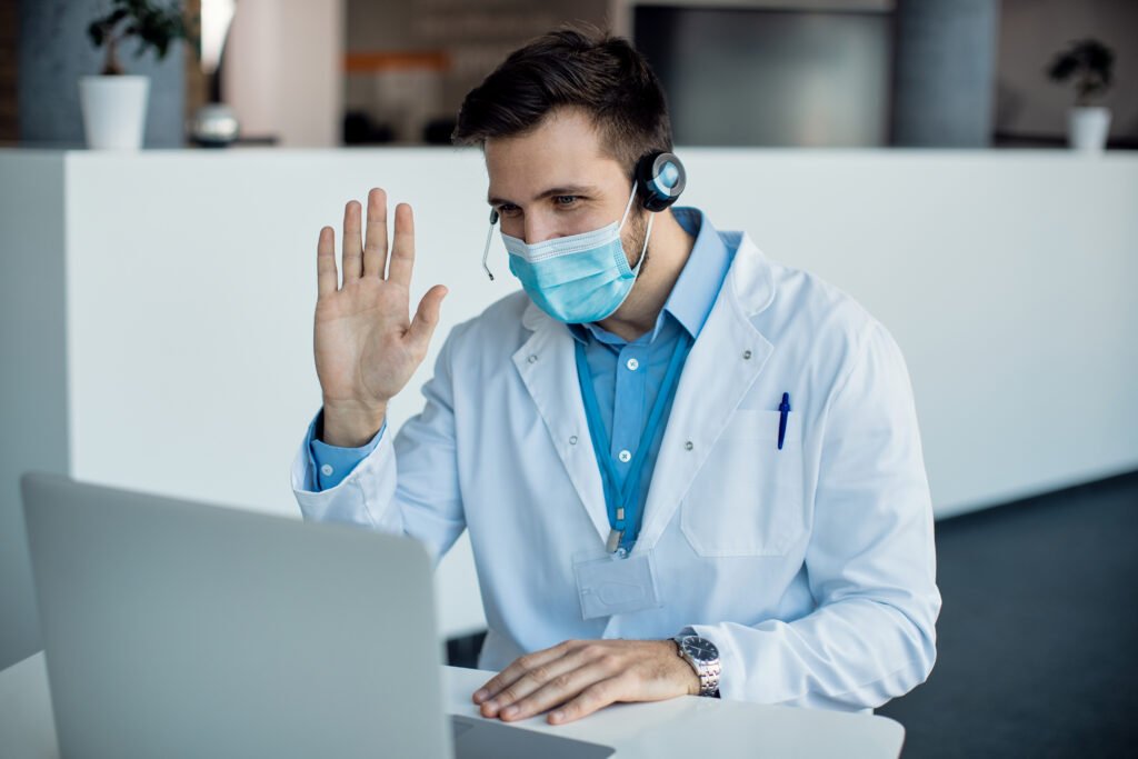 Home male doctor waving during a video call over laptop at medical c